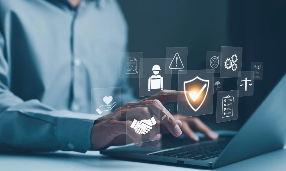 Person using a laptop with floating digital icons for safety, corporate security compliance, and operational management, highlighting a shield with a checkmark