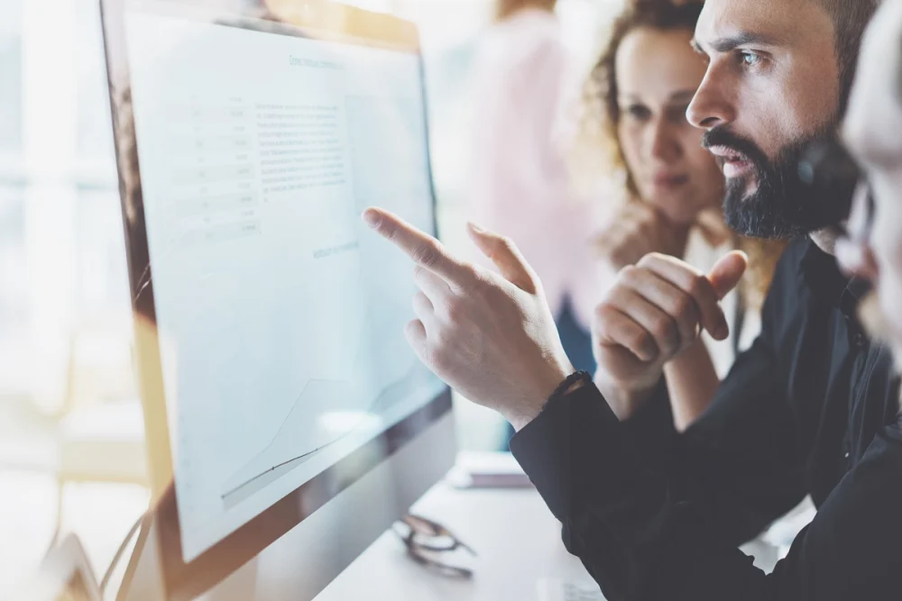 Close-up of colleagues collaborating at a computer on corporate compliance plans, with one person pointing at data and charts displayed on a large monitor